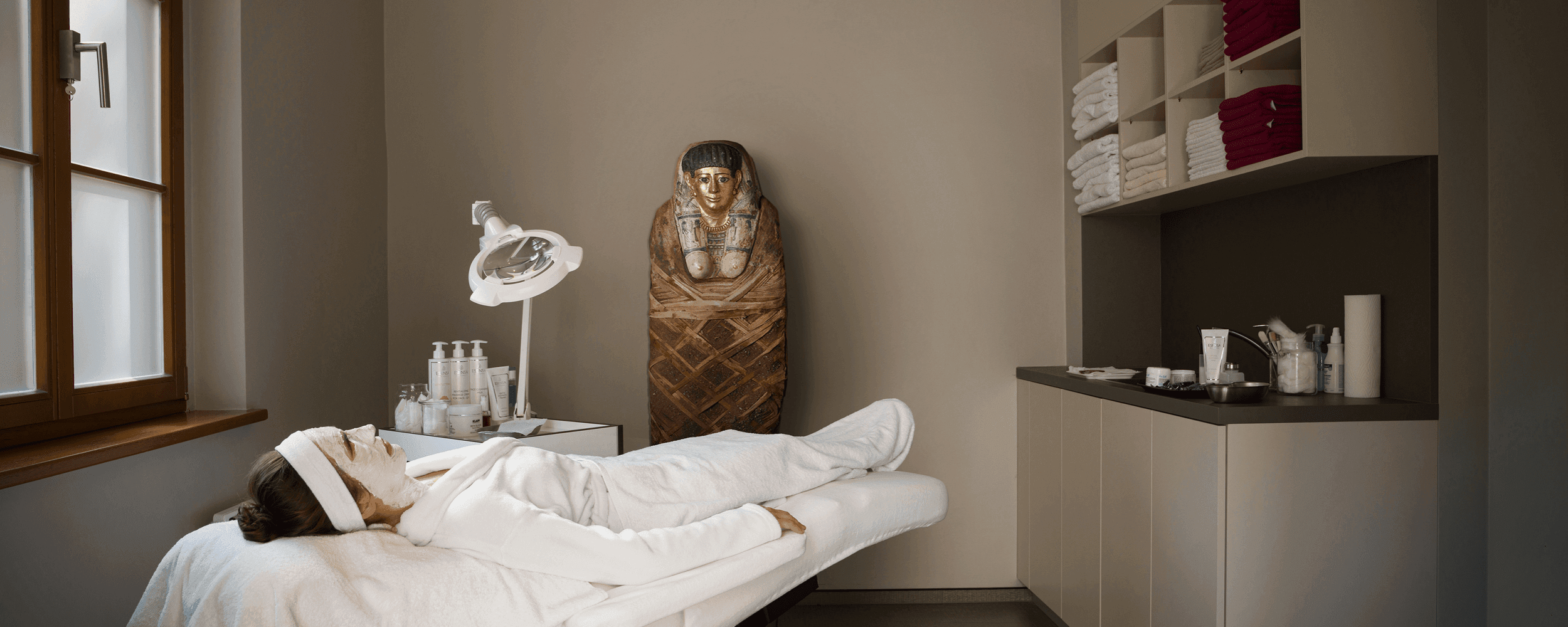 Person liegt mit Gesichtsmaske entspannt auf einer Spa-Liege; im Raum stehen Pflegeprodukte und eine ägyptische Mumienfigur Person liegt mit Gesichtsmaske entspannt auf einer Spa-Liege; im Raum stehen Pflegeprodukte und eine ägyptische Mumienfigur